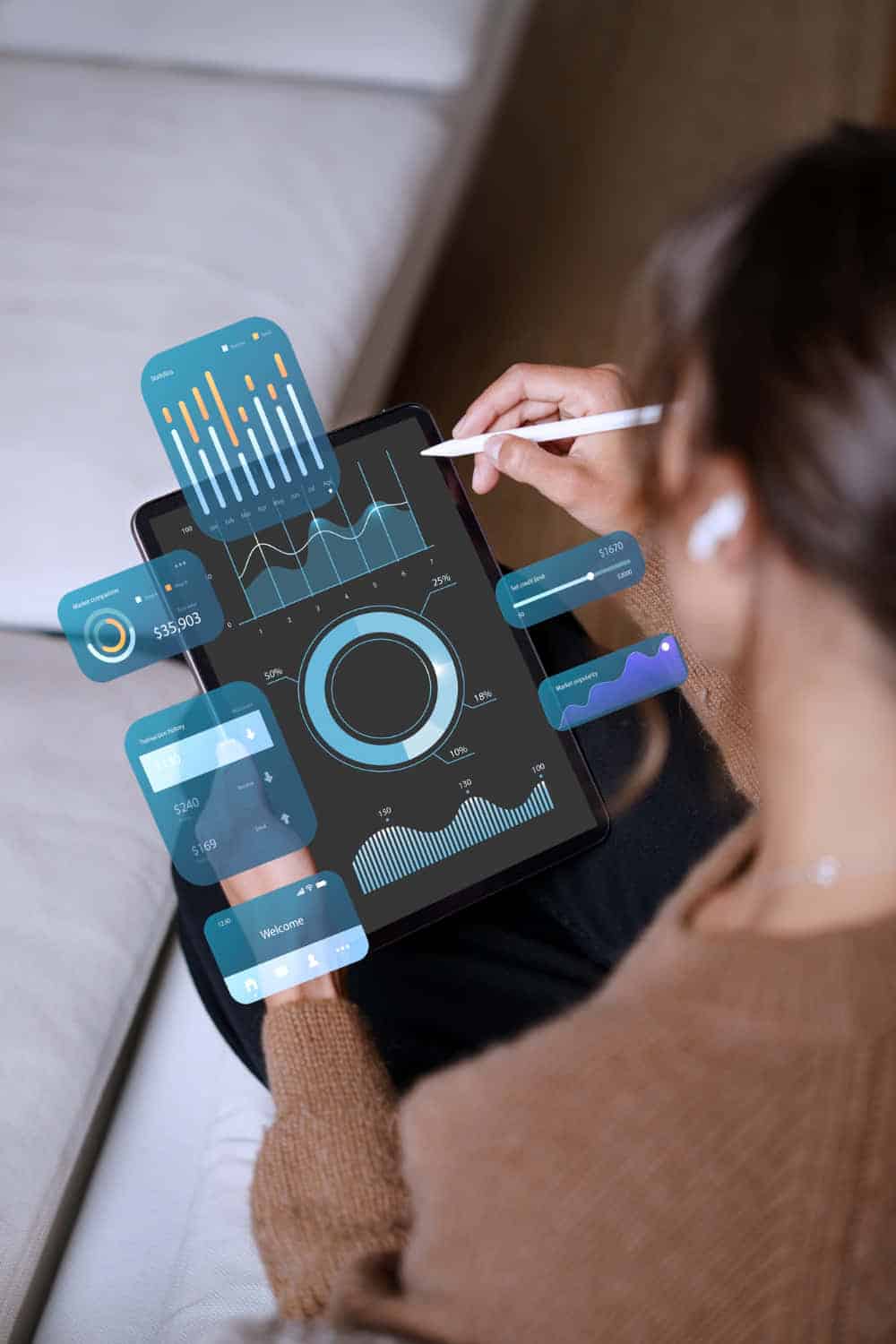 Vue de dos d'une personne assise sur un canapé, utilisant un stylet sur une tablette tactile affichant des graphiques analytiques (courbes, barres, diagrammes circulaires) avec des éléments holographiques superposés en bleu et orange, comme des données financières et des indicateurs de performance, dans un environnement cosy.