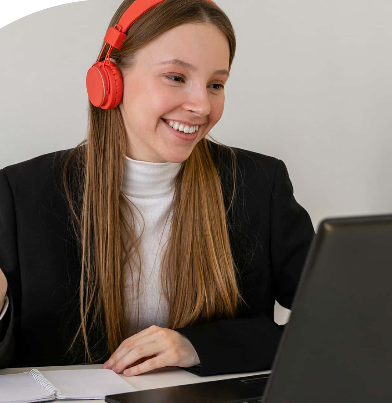 Jeune femme souriante portant un casque audio rouge vif, concentrée et heureuse devant son ordinateur portable ouvert, veste noire et col roulé blanc, carnet à spirale posé sur le bureau.
