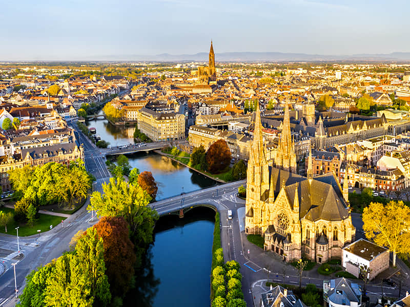 Vue aérienne de Strasbourg au soleil couchant, avec la cathédrale Notre-Dame et ses flèches gothiques dominant la vieille ville, la rivière Ill traversée par plusieurs ponts, des bâtiments historiques aux toits rouges et des arbres aux feuillages d’automne.